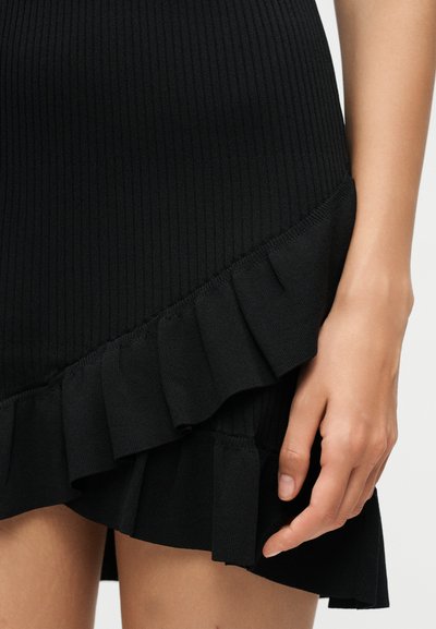 Κοντινό πλάνο μιας μαύρης πλεκτής φούστας με στρωμένο βολάν στο τελείωμα, που δείχνει ένα χέρι να ακουμπά απαλά στο μηρό.