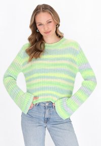Junge Frau mit welligem blonden Haar, die einen gr�ün-blauen gestreiften Ripppullover und hellblaue hoch taillierte Jeans trägt, mit den Händen in den Taschen.