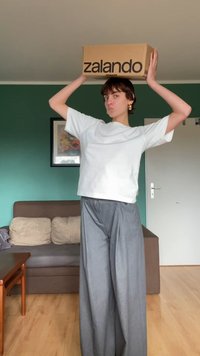 Caixa marrom da Zalando segurada acima da cabeça por uma pessoa que veste uma blusa branca de mangas curtas e calças largas cinzas. Interior com parede verde e sofá.