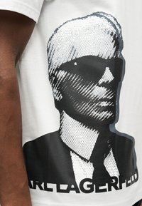 Μαύρο και λευκό στυλιζαρισμένο γραφικό ενός ατόμου που φορά γυαλιά ηλίου και κοστούμι, τυπωμένο σε λευκό T-shirt.