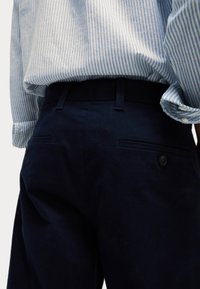 Pantaloni blu navy con una texture liscia, dotati di due tasche posteriori e chiusura con bottone. Abbinati a una camicia a righe azzurre.
