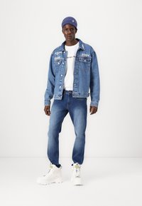 Chaqueta de denim con detalles bordados en azul claro, combinada con una camiseta blanca y jeans de denim azul. Llevada con zapatillas blancas y un gorro azul.