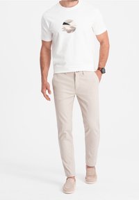 Mężczyzna ubrany w białą koszulkę z grafiką, beżowe spodnie slim-fit, beżowe buty wsuwane oraz czarny zegarek na nadgarstku, stoi z jedną ręką w kieszeni.