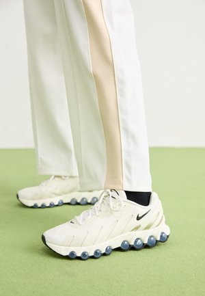 Baskets blanches Nike avec semelle en forme de bulles, portées avec un pantalon blanc orné d'une bande latérale beige, debout sur un sol vert.