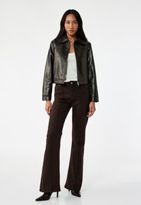 Giacca nera in pelle cropped sopra una maglietta bianca, abbinata a jeans svasati marrone scuro con dettagli di cucitura e tacchi a punta neri.