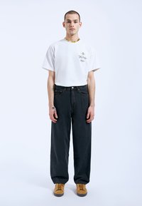 White short-sleeve T-shirt with graphic design, paired with loose-fitting dark denim jeans and brown suede shoes, displaying a relaxed silhouette.