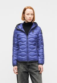 Jeune personne aux cheveux courts roux portant une veste matelassée bleue par-dessus un col roulé noir et un jean noir délavé.