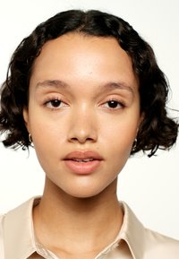 Gros plan sur une personne aux cheveux frisés et sombres, portant une chemise beige clair. Sa peau est lisse avec un maquillage naturel ; les lèvres sont légèrement colorées.