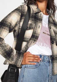 Koszula flanelowa w kratę w kolorach kremowym, czarnym i szarym; zapinana na guziki, z dwoma kieszeniami na piersi, noszona na koszulce z grafiką i wysokim stanem jeansów.