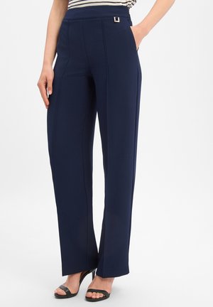Vrouw draagt marineblauwe, recht gesneden broek met hoge taille en zijzakken, en zwarte open-toe hakken, staand tegen een witte achtergrond.