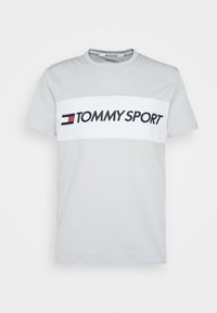 Camiseta de algodón gris claro con una banda horizontal blanca con "TOMMY SPORT" en negro, acentuada por un logo en rojo y azul marino. Manga corta.