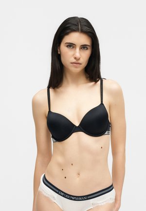 Γυναίκα με μακριά σκούρα μαλλιά, που φοράει μαύρο σουτιέν και λευκά εσώρουχα με δαντελένιο τελείωμα και το λογότυπο Emporio Armani στη ζώνη, πάνω σε λευκό φόντο.
