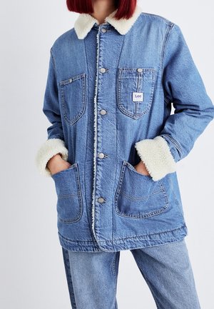 Osoba nosząca niebieską kurtkę dżinsową z białym kołnierzem i mankietami sherpa, ręce w przednich kieszeniach, w zestawie z dopasowanymi dżinsami.