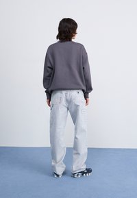 Grijze oversized sweatshirt, lichtblauwe cargo jeans met zakken en donkere sportschoenen. Achtergrond is effen wit met een blauwe vloerbedekking.