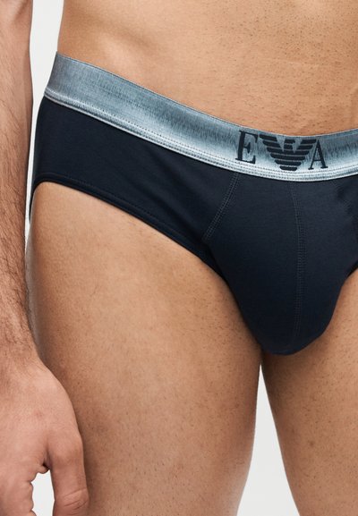 Άνδρας που φοράει μαύρα σλιπ με ασημένια μέση με το λογότυπο "EA" και έμβλημα αετού, εμφανιζόμενος από τη μέση μέχρι το μέσο του μηρού.