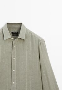 Camicia di lino verde chiaro con chiusura a bottoni, maniche lunghe e righe bianche. Presenta un colletto classico e piccoli bottoni.