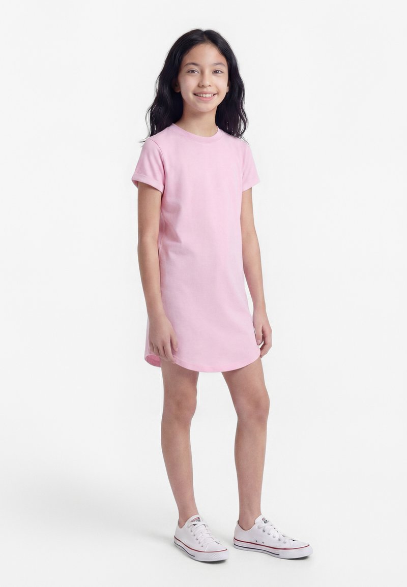 Usmievavé dievča s dlhými tmavými vlasmi, ktoré má na sebe ružové šaty s krátkym rukávom a biele tenisky, stojí pred bielym pozadím.