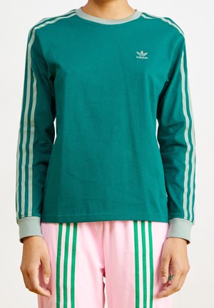 Personne portant un haut à manches longues Adidas turquoise avec des rayures bleu clair et un pantalon rose avec des rayures vertes, visible du cou jusqu'aux hanches.