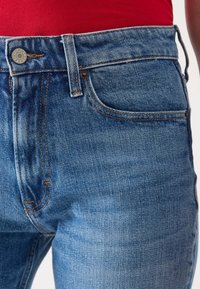 Primer plano de la cintura de unos pantalones de mezclilla azul y el bolsillo frontal con costuras visibles y un botón de metal, usados con una camiseta roja.