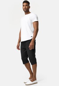 Camiseta blanca, pantalones cortos negros hasta la rodilla y zapatillas blancas sin cordones. La camiseta presenta un diseño simple con una pequeña etiqueta en el dobladillo.