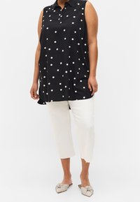 Schwarzes ärmelloses Oberteil mit weißen Punkten, spitzem Kragen und lockerer Passform, kombiniert mit cropped weißen Hosen und gestreiften Slippern.