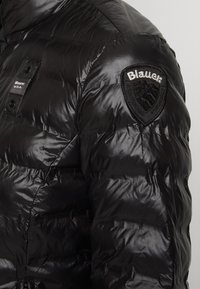 Czarna kurtka puchowa o błyszczącym wykończeniu. Posiada materiałową naszywkę z napisem "Blauer" oraz wzorem tarczy na lewym ramieniu.