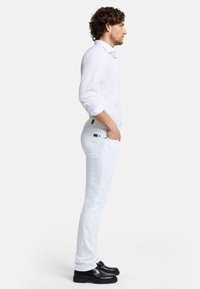 Chemise blanche à boutons, avec des manches retroussées, associée à un pantalon blanc et des chaussures noires. La tenue présente un design élégant et moderne.
