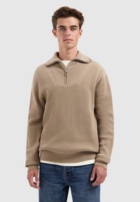 Beige half-zip pullover in ribbelstructuur met een kraag. In combinatie met blauwe jeans, met daaronder een gelaagd wit shirt.