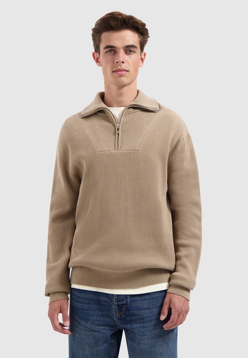 Beige half-zip pullover in ribbelstructuur met een kraag. In combinatie met blauwe jeans, met daaronder een gelaagd wit shirt.