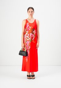 Mujer con un vestido midi sin mangas de flores rojas, sosteniendo un bolso negro con asas doradas y usando sandalias de tacón negro.