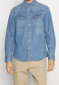 Ljust blå denimskjorta med långa ärmar, knappad krage, två bröstfickor och enkel-knappmanschetter; har en något rundad nederkant.