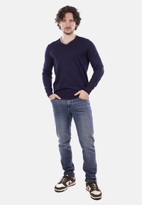 Pullover blu navy a collo a V realizzato in tessuto lavorato, abbinato a jeans leggeri e sneaker marroni. Il modello si trova in piedi con le mani nelle tasche su uno sfondo neutro.