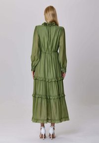 Femme portant une longue robe olive claire et transparente avec des volants et des manches longues, de dos, associée à des sandales à talons blancs.