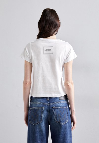Hvit avkortet t-skjorte med korte ermer har et rektangulært svart trykk på ryggen. Kombinert med blå jeans, som har v-formede lommer.