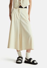 Krémové kalhoty širokého st řihu vyrobené z měkké látky, s vysokým pasem, předními kapsami a hladkým povrchem, doplněné černými sandály a bílými ponožkami.