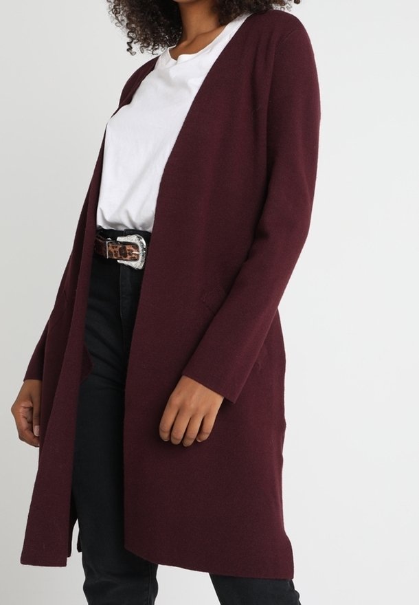 Personne portant un long cardigan bordeaux, un t-shirt blanc, un pantalon noir et une ceinture à imprimé léopard avec une boucle argentée.