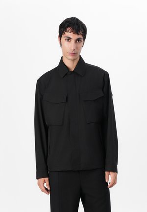 Jachetă neagră cu guler, două buzunare frontale și o croială lejeră. Confecționată dintr-un material neted cu un design minimalist.