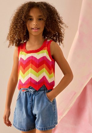 Kind mit lockigem Haar trägt ein ärmelloses, farbenfrohes Top mit Zickzackmuster und blaue Jeansshorts, steht mit einer Hand in der Tasche.