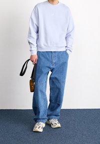 Light purple sweatshirt, loose-fitting blue denim jeans with patchwork stitching, and chunky white sneakers, holding a small brown bag.