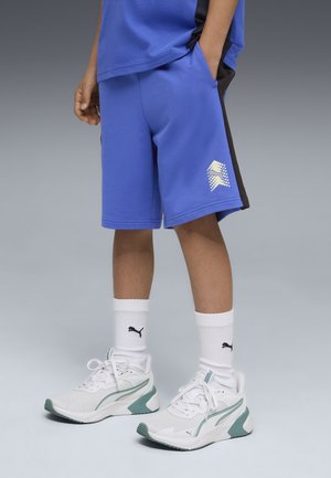 Niño con pantalones cortos azules, calcetines blancos de Puma y zapatillas deportivas blancas con detalles verdes, de pie con una mano en el bolsillo.