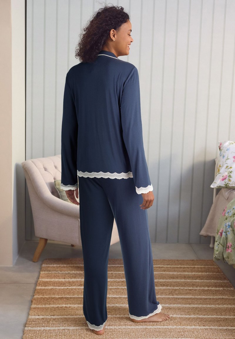 Marineblaues Pyjama-Set mit langärmeligem Oberteil und Hose, versehen mit weißer Spitzenverzierung an Bündchen und Saum. Weicher Stoff, lockere Passform.