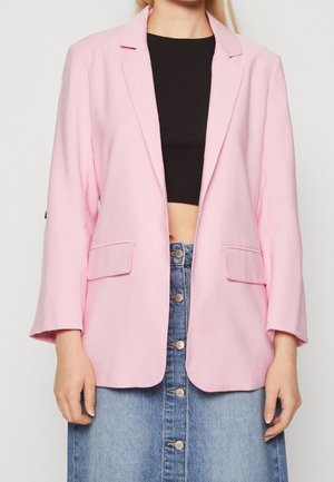Vrouw draagt een lichtroze blazer over een zwarte cropped top en een hooggetailleerde spijkerrok met knopen tegen een effen achtergrond.