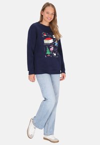 Sudadera azul marino con gráficos coloridos de temática navideña. Combinada con vaqueros azul claro y zapatillas blancas, mostrando un estilo casual.