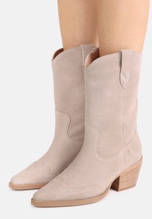 Lys beige ruskind cowboystøvler midt på læggen med spidse tæer, træblokhæl og dekorativ syning, båret på ben.