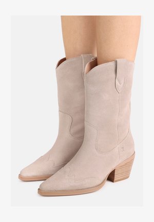 Hellbeige Wildleder-Cowboystiefel bis zur Mitte der Wade mit spitzen Zehen, hölzernen Blockabsätzen und dekorativen Nähten, getragen an den Beinen.