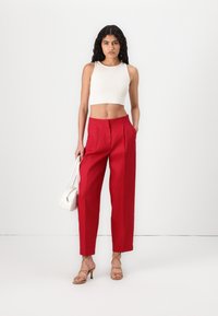 Pantalons taille haute rouges avec des plis, associés à un débardeur beige court. Les accessoires comprennent un sac à main blanc et des chaussures à talons en cuir brun.