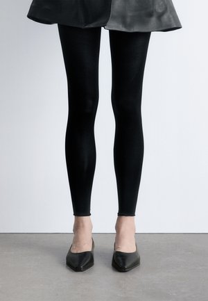Jambes portant des leggings noirs associés à une jupe en cuir noire et des talons pointus noirs, debout sur un sol gris.