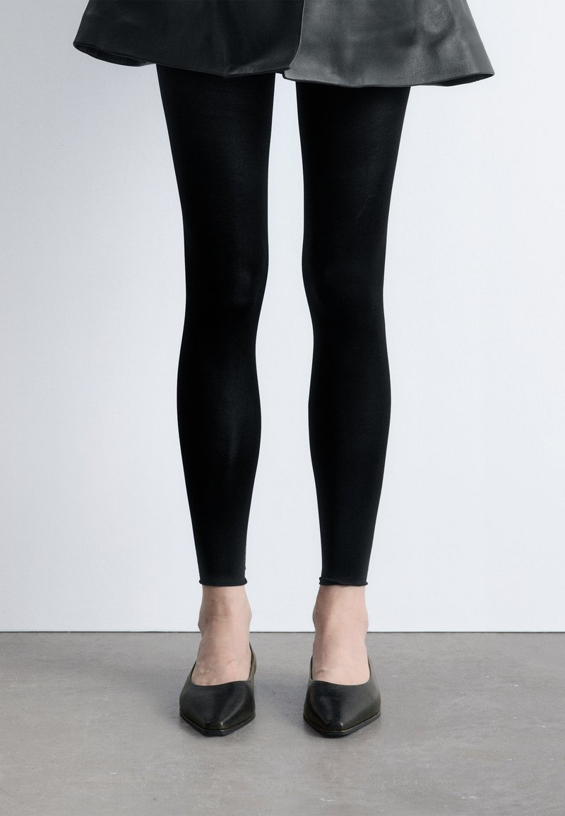 Jambes portant des leggings noirs associés à une jupe en cuir noire et des talons pointus noirs, debout sur un sol gris.