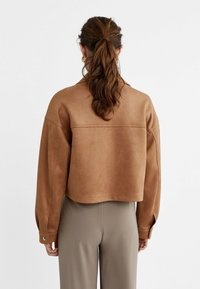 Femme avec une longue queue de cheval brune portant une veste en suède beige courte et un pantalon taille haute taupe, se tenant devant un fond blanc uni.