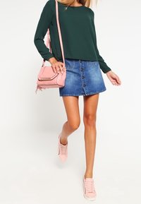 Un top verde a maniche lunghe, una gonna in denim con bottoni frontali, una borsa a tracolla rosa e delle sneakers rosa chiaro creano un outfit casual con colori e materiali distintivi.